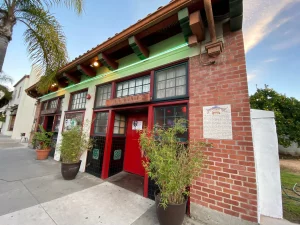 Pickle Room red door with palm tree, plants, and sidewalk in front.