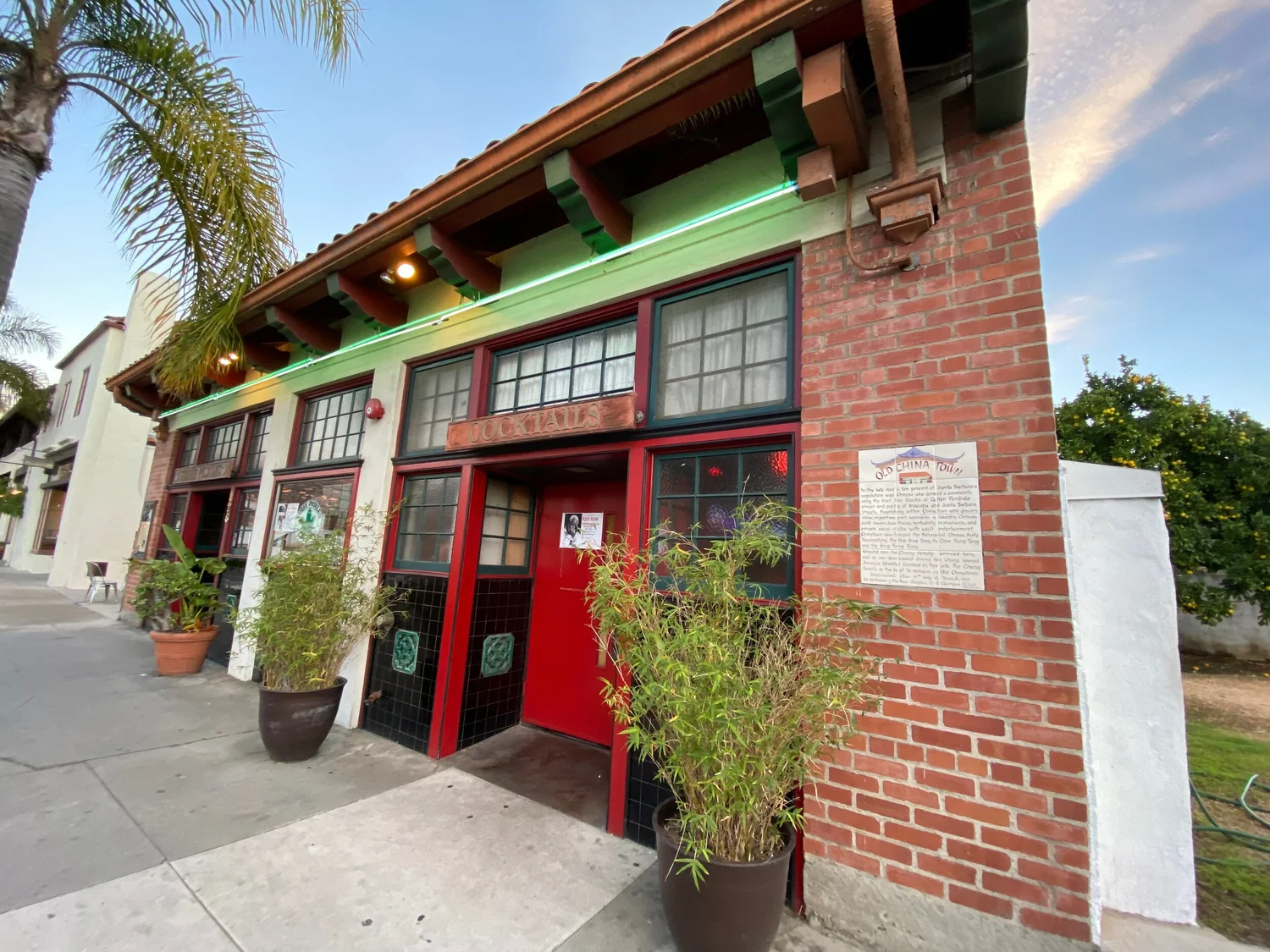 Pickle Room red door with palm tree, plants, and sidewalk in front.